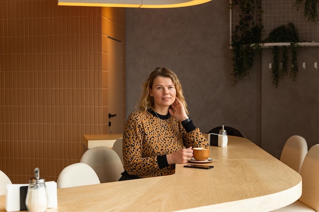 Foto een jonge blonde vrouw zit in een café in beige tinten koffie te drinken, scrollend aan de telefoon en te lachen