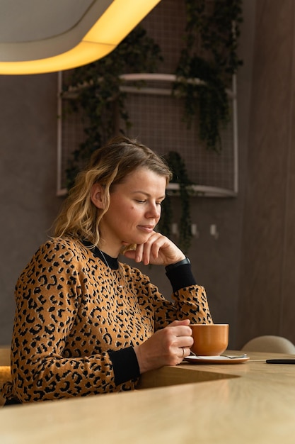 Foto een jonge blonde vrouw zit in een café in beige tinten koffie te drinken, scrollend aan de telefoon en te lachen