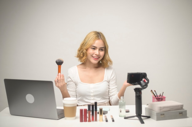 Een jonge blonde vrouwelijke ondernemer die met laptop werkt, presenteert cosmetische producten tijdens online livestream over witte achtergrondstudio, die online en beauty blogger-concept verkoopt