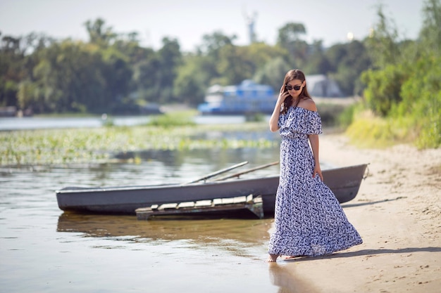 Een jonge brunette in een lange jurk staat bij de boot aan de oevers van de rivier Copy space