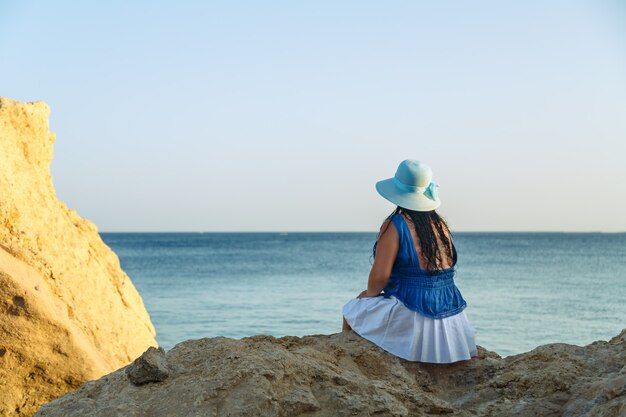 Een jonge brunette vrouw in een witte rok en blauwe hoed aan de kust zit op de rotsen met haar rug