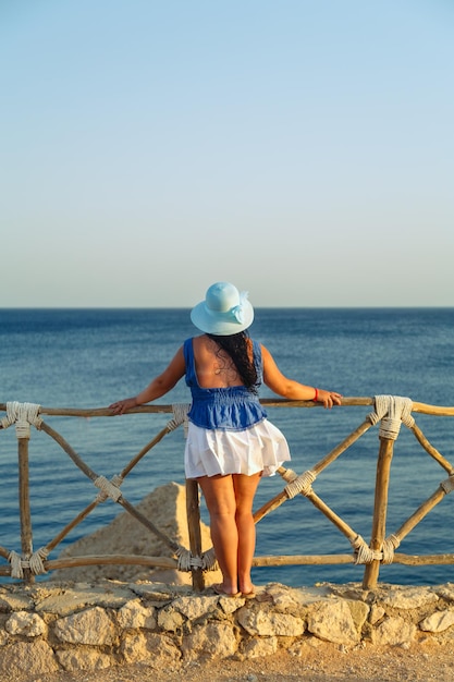 Een jonge brunette vrouw in een witte rok en een blauwe hoed aan de kust bewondert de zee