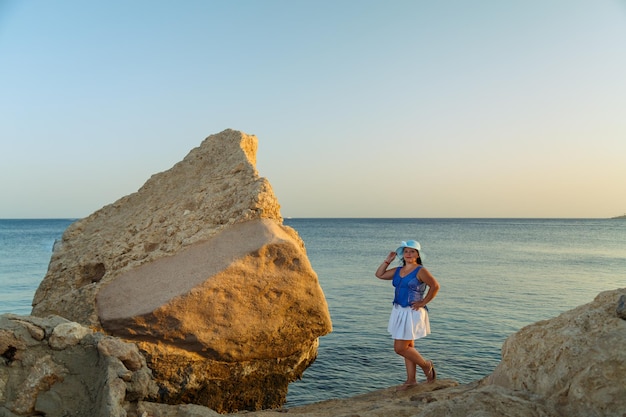 Een jonge brunette vrouw in een witte rok en een zonnehoed aan de kust op een rots bewondert de natuur.
