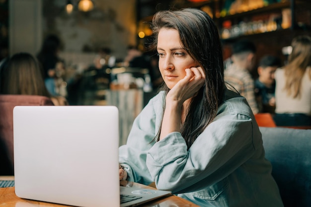 Een jonge brunette vrouw zit in een café en werkt aan een laptop.