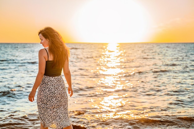 Een jonge charmante vrouw in een rok staat aan de kust voor een feloranje zonsondergang