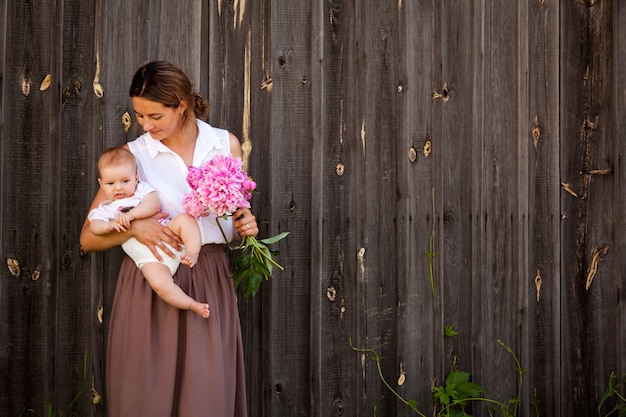 Een jonge donkerharige vrouw in een wit overhemd in een beige rok houdt een kleine baby in haar hand en een mooi boeket roze pioenrozen op de achtergrond van een houten hek