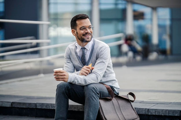 Een jonge, elegante man zit buiten op een trap koffie te drinken en een e-sigaret te roken