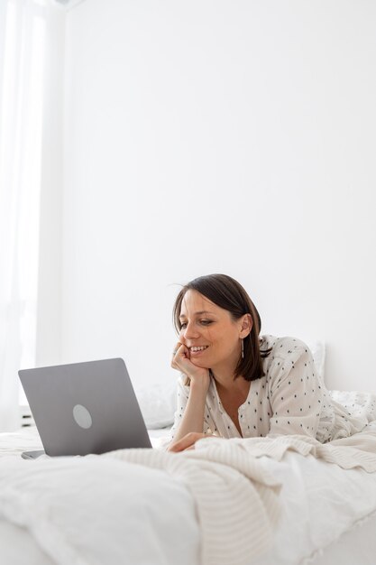 Een jonge en mooie Europese vrouw liggend op een bed in een witte slaapkamer met een laptop