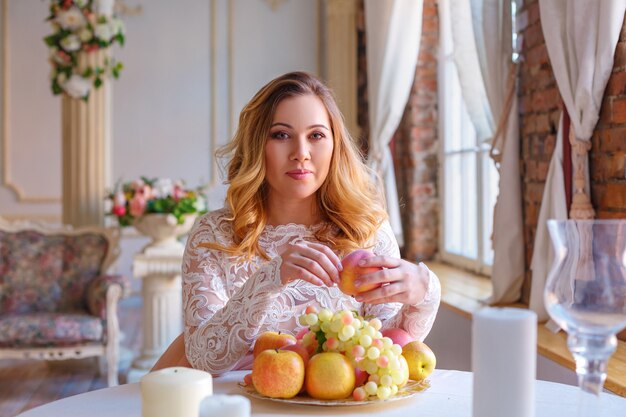 Een jonge en mooie vrouw eet een perzik aan een tafel vol met fruit in het binnenland