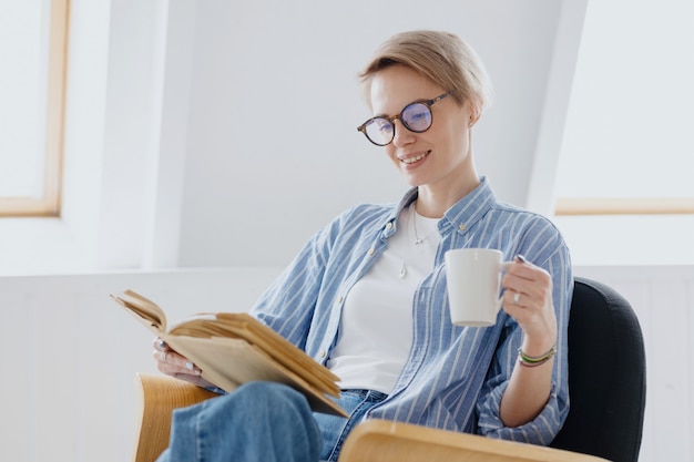 Een jonge Europese vrouw met kort blond haar drinkt koffie of thee en leest een boek