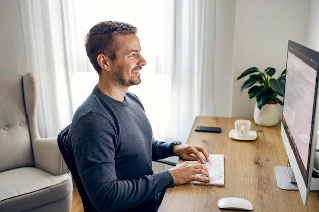 Een jonge freelancer zit op het thuiskantoor en typt een e-mail terwijl hij naar de monitor glimlacht