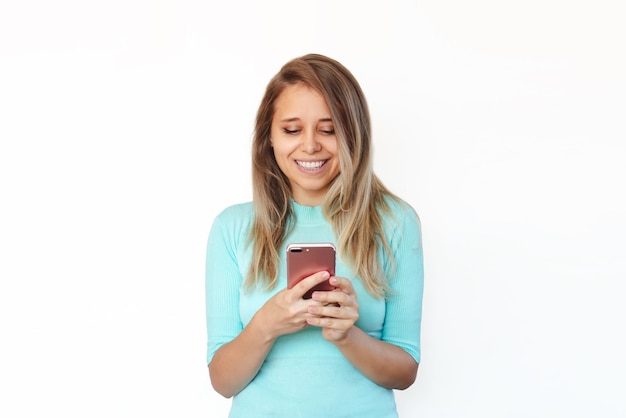 Een jonge, gelukkige blonde vrouw glimlacht en houdt een mobiele telefoon vast terwijl ze naar het scherm kijkt