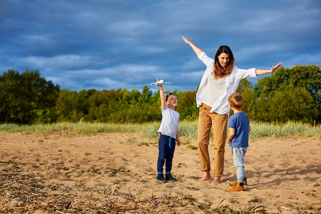 Een jonge gelukkige moeder met twee jongens die op het zand in de buurt van het bos spelen, in handen van een van hen een model van een civiel vliegtuig en een hoed op zijn hoofd