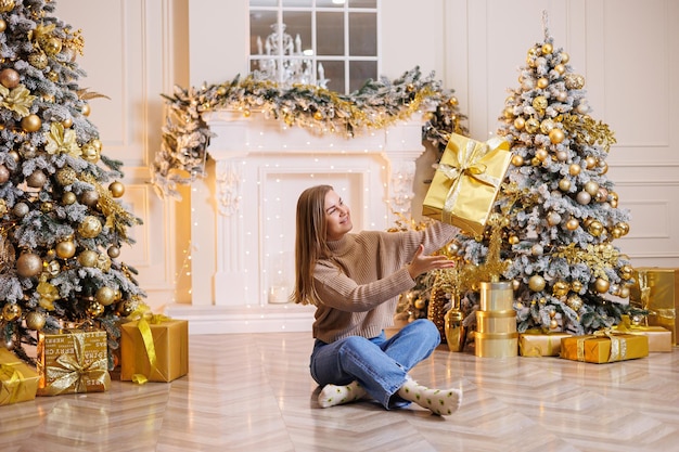 Een jonge gelukkige vrouw zit met een nieuwjaarscadeau-kerstboom en een open haard met feestelijke slingers nieuwjaarsstemming