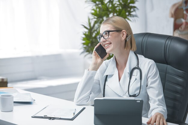 Een jonge gespecialiseerde arts werkt aan een bureau in zijn heldere kantoor en praat aan de telefoon