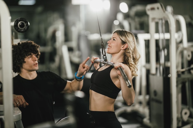 Een jonge gespierde vrouw traint op een machine die door een coach in de sportschool wordt ondersteund.