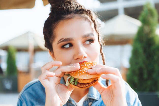 Een jonge glamoureuze vrouw met dreadlocks en rode lippenstift zit en eet een hamburger in een straatcafé, het concept van het eten van langdurige lippenstift