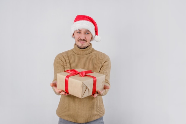 Een jonge glimlachende blanke man met een kerstmuts die een cadeau uithoudt en naar de camera kijkt op een witte geïsoleerde achtergrond