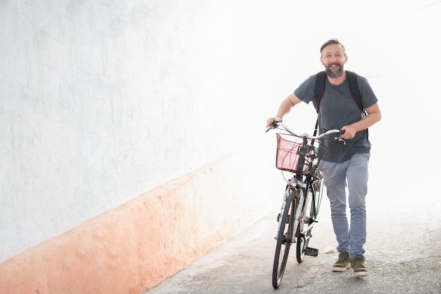 Foto een jonge glimlachende stijlvolle baardige hipster man met een rugzak die een retro fiets duwt terwijl hij op straat loopt