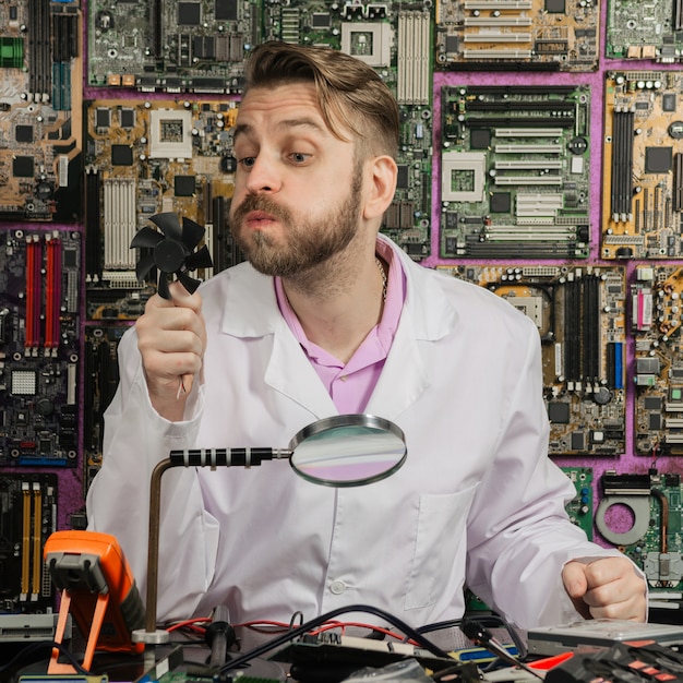 Een jonge grappige elektronica-ingenieur blaast op een computer fansitting aan zijn laboratoriumtafel