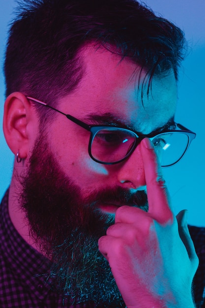 Een jonge hipster man met blauw licht bril wegkijken naar camera met een ernstig gezicht op blauwe en roze kleuren terwijl het aanraken van de bril