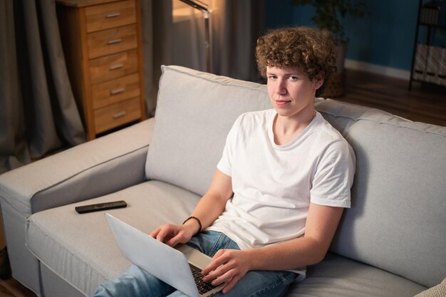 Een jonge jongen met krullend haar zit op de bank in de woonkamer met zijn laptop op schoot