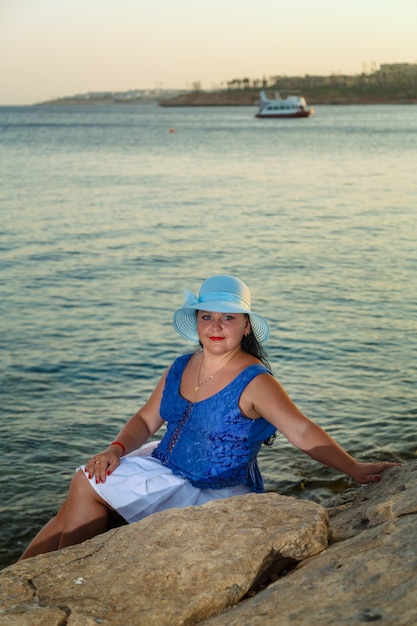 Een jonge Joodse vrouw in een witte rok en een zonnehoed aan de kust zit op een rots. Verticale foto.