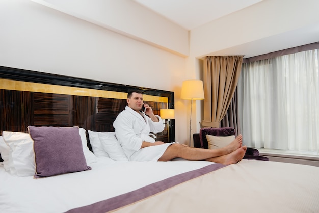 Een jonge kerel in een witte laboratoriumjas zit op het bed en praat aan de telefoon in zijn hotelkamer.
