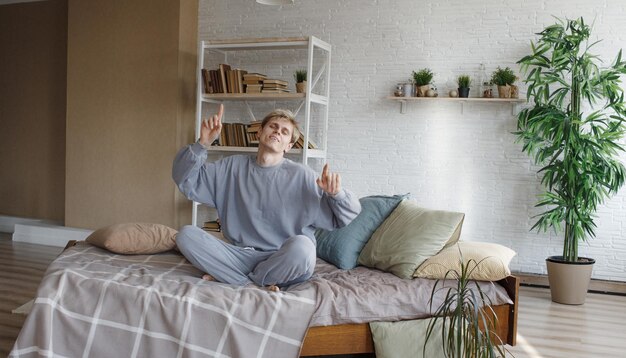 Een jonge kerel in grijze pyjama danst zittend op het bed met zijn handen omhoog