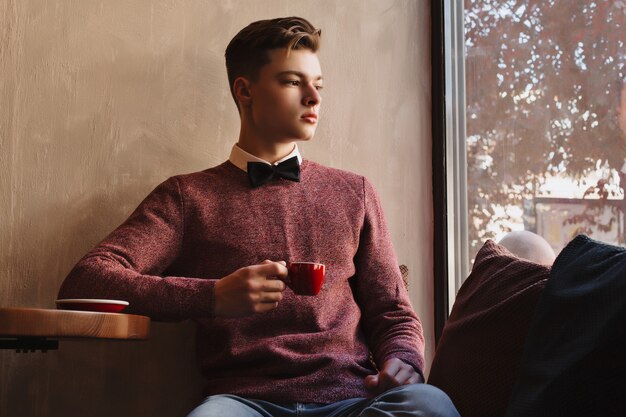 Een jonge kerel met een vlinderdas droomt van de toekomst en drinkt koffie uit een rode mok