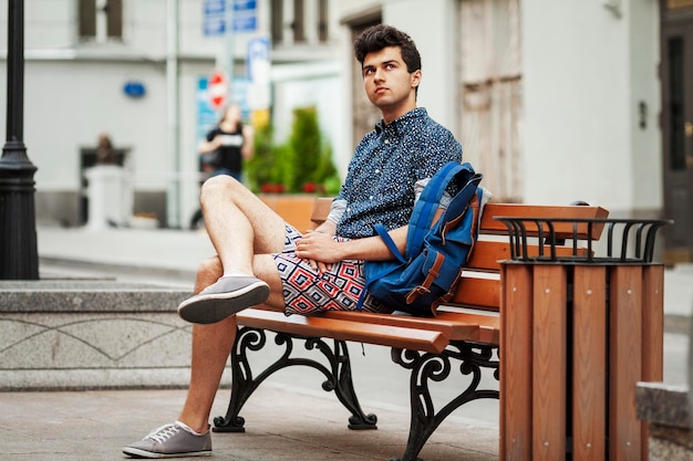 Een jonge kerel zit op een bankje in de stad brunette in korte broek en een blauw shirt met een rugzak