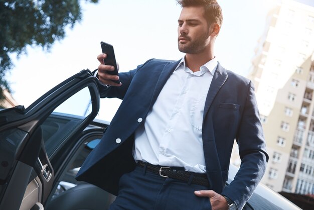 Een jonge knappe man in een pak komt uit de auto en gebruikt een smartphone