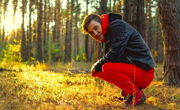 Foto een jonge knappe man in een rood warm pak en een leren jas zit op je hurken in het bos