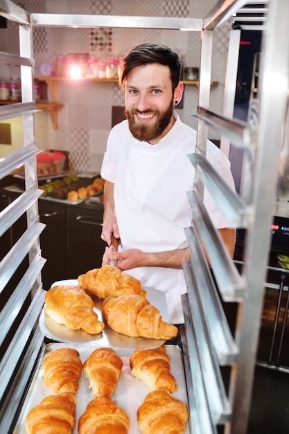 Een jonge knappe mannelijke bakker houdt een dienblad met Franse croissants voor een bakkerij en glimlacht.