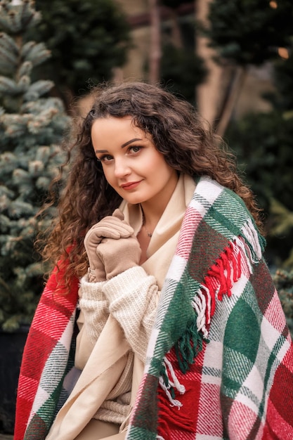 Een jonge krullende vrouw gewikkeld in een warme plaid in een cel zit te wachten op het nieuwe jaar het meisje op de achtergrond van groene kerstbomen op straat vakantiestemming