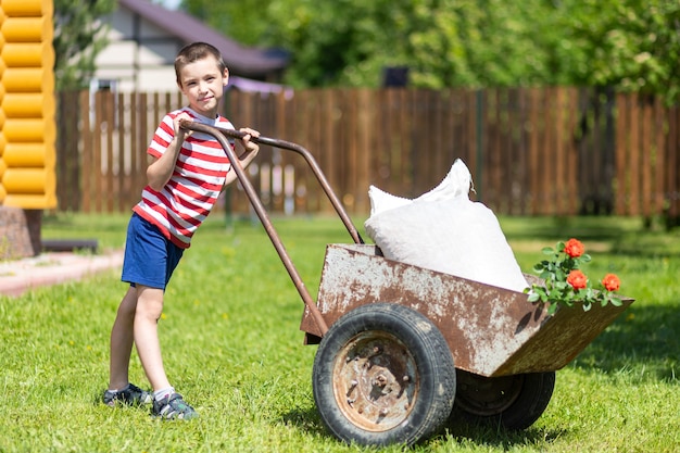 een jonge lachende jongen duwt een kruiwagen over de tuin. assistent jongen in t-shirt en korte broek