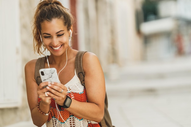 Een jonge lachende vrouw die muziek luistert en haar smartphone gebruikt terwijl ze tijd doorbrengt op vakantie in een mediterrane stad.