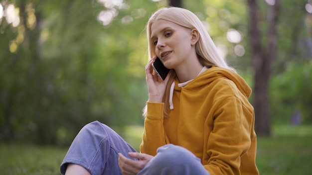 Een jonge lachende vrouw zit in een groene weide en praat aan de telefoon. De vreugde van een telefoongesprek, prettige communicatie. Camping. Close-up, 4K UHD.