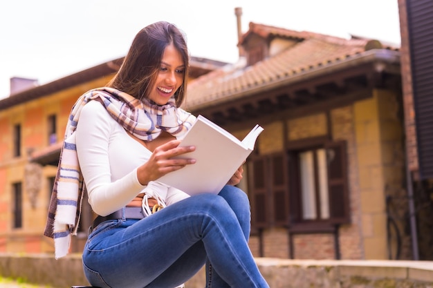 Een jonge Latijnse vrouw met een witte t-shirtjeans en een sjaal die geniet van en zich voorstelt een boek te lezen in de oude stad Kijkend naar de camera en glimlachen