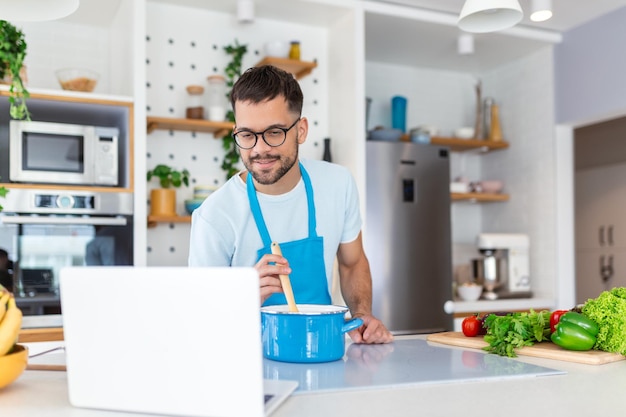 Een jonge man brengt een dag thuis door en bereidt voedsel van groenten in de keuken een man in huiskleren en met een baard gebruikt een laptop om online koken te leren