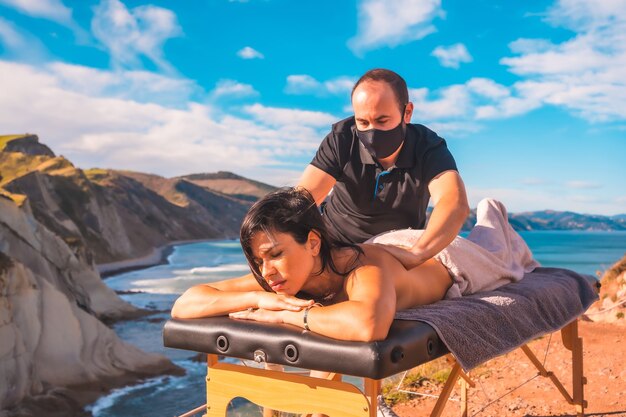 Een jonge man die een massage uitvoert in de natuur aan de kust bij de zee, een droom die uitkomt, masseuse met gezichtsmasker in de coronavirus-pandemie