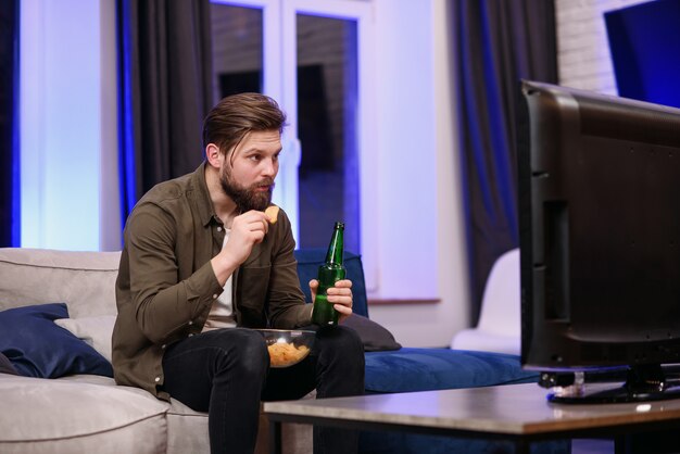 Een jonge man die op de bank in de woonkamer zit, is geschokt door een film op de tv 's nachts.