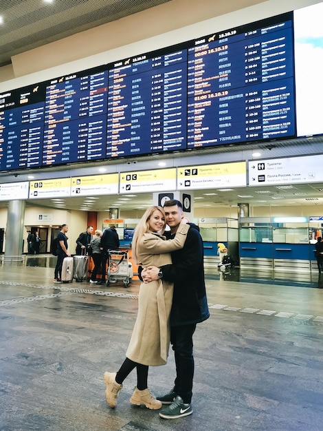 Een jonge man en vrouw omhelzen elkaar op de luchthaven voor het vluchtinformatiebord
