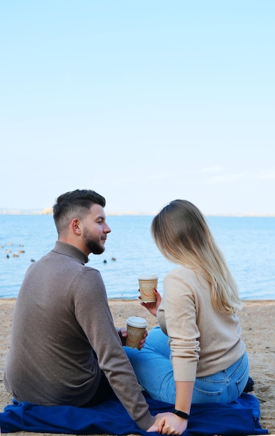 Een jonge man en vrouw zitten op het strand met hun rug naar de camera met glazen koffie in hun handen De man kijkt liefdevol naar het meisje