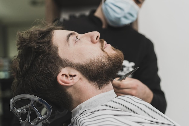 Een jonge man fixeert zijn baard in de kapperszaak De kapper draagt een beschermend masker