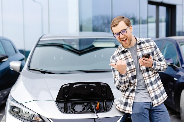 Een jonge man gebruikt een mobiele telefoon terwijl hij op zijn elektrische auto leunt terwijl de auto wordt opgeladen