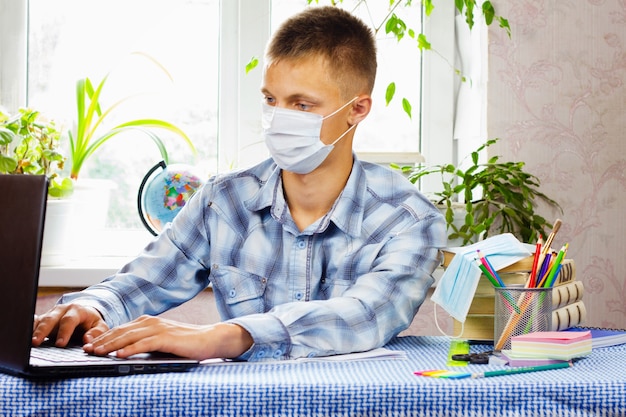 Een jonge man, gekleed in een geruit overhemd en een steriel masker, zit aan een tafel en kijkt naar een laptop. Onderwijs concept. Slechte epidemiologische situatie.