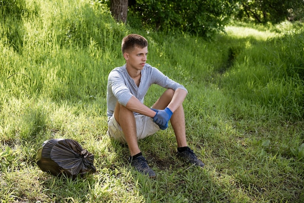 Een jonge man heeft het park vrijwillig schoongemaakt van vuilnis en zit op het gras met handschoenen die rusten na het werk