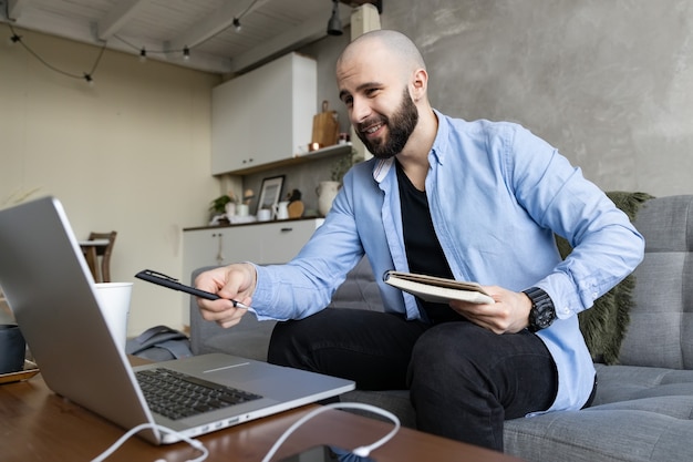 Een jonge man in een blauw shirt en zwarte spijkerbroek studeert online vanuit huis met behulp van een computer en een dagboek voor aantekeningen. Concept van werk en studie online