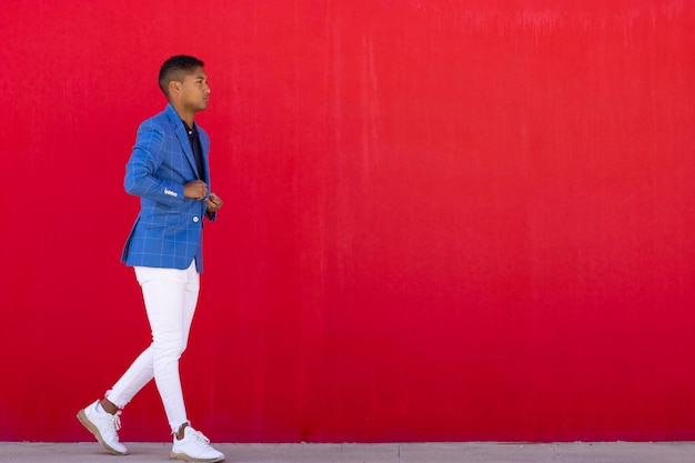 Een jonge man in een blauwe jas en een witte broek die tegen een rode muur loopt